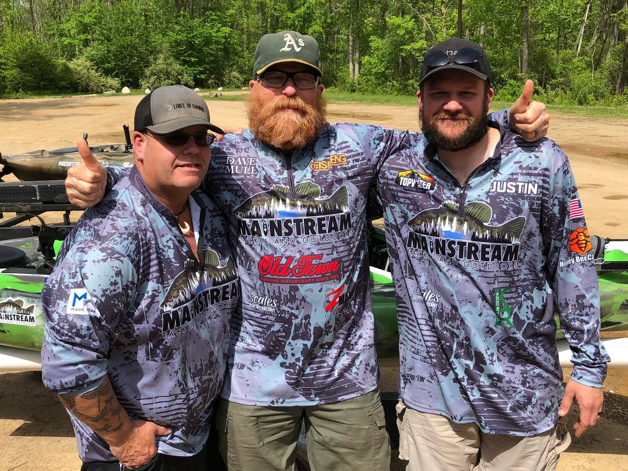Three men in matching fishing shirts giving thumbs up outdoors.