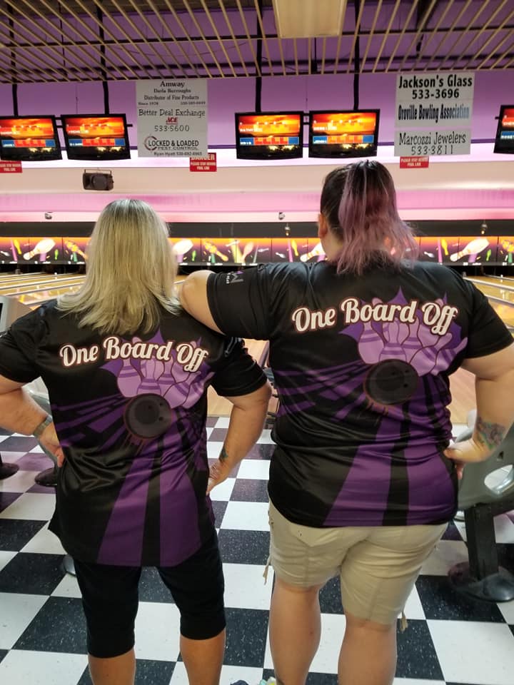 Two women wearing &#39;One Board Off&#39; shirts at a bowling alley.