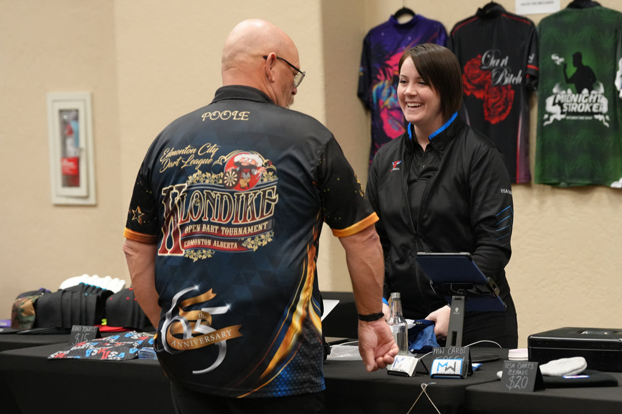 Customer interacting with a Magic Wear salesperson at a booth with merchandise on display during USA Dart Classic.